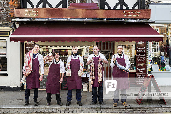 Gruppe von Männern  Metzger und Fischhändler  mit Schürzen bekleidet  stehen auf dem Bürgersteig vor einer Metzgerei  halten Fleisch- und Fischstücke in der Hand und schauen in die Kamera.