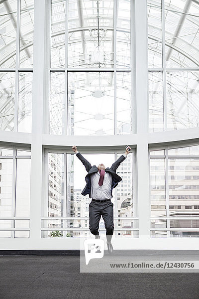 A businessman jumping for joy in front of a large window in a convention centre lobby.