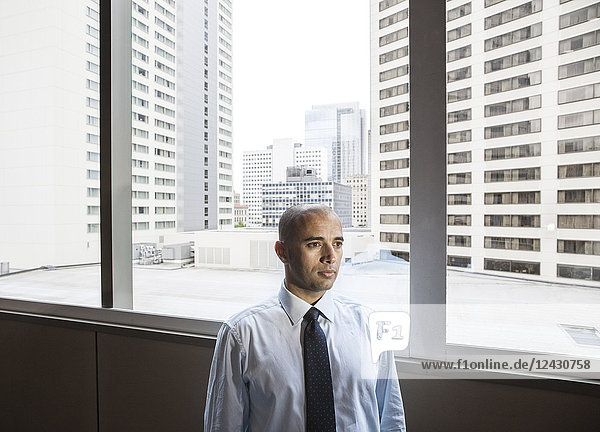 Ein Geschäftsmann aus dem Nahen Osten steht neben einem Fenster  das auf eine Stadtlandschaft mit hohen Bürogebäuden blickt.