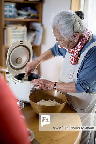 Ältere Frau  die an einem Tisch in einer Küche steht und Sushi-Reis vom Reiskocher in eine Schüssel löffelt.