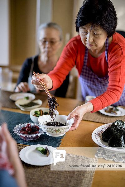 Woman wearing apron standing at kitchen table holding bowl serving food with chopsticks.