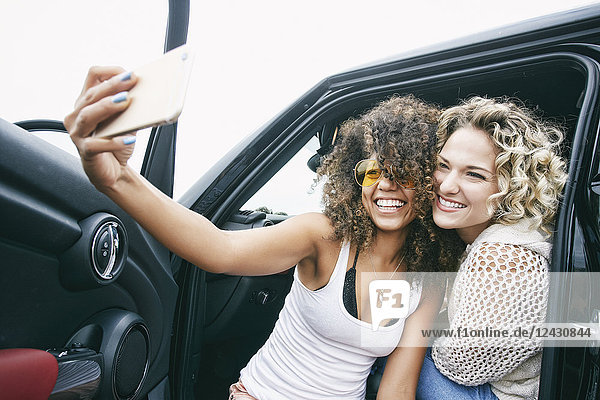 Porträt von zwei lächelnden jungen Frauen mit blonden und braunen Locken  die im Auto sitzen und sich mit dem Handy selbstständig machen.