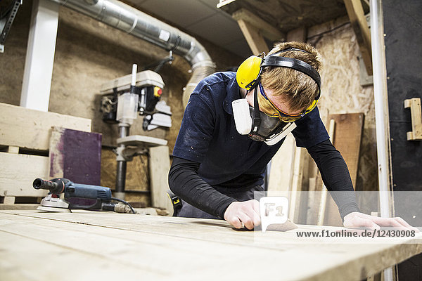 Mann mit Gehörschutz  Schutzbrille und Staubmaske steht in einem Lagerhaus und arbeitet an einem Stück Holz.
