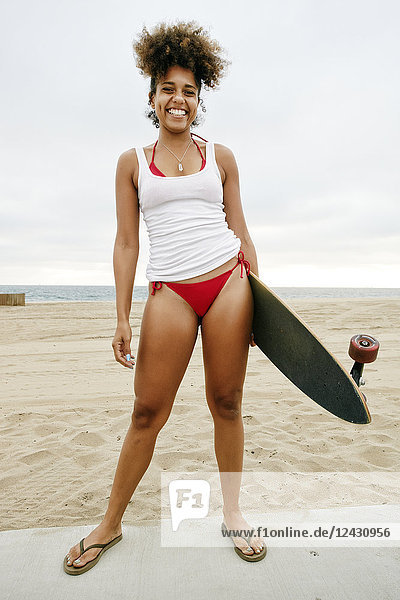 Young woman with curly brown hair wearing white vest and red bikini standing on sandy beach  holding skateboard.