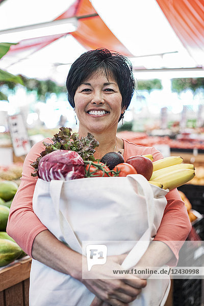 Lächelnde Frau steht in einem Lebensmittel- und Gemüsemarkt und hält eine Einkaufstasche mit frischen Produkten wie Bananen  Tomaten und Kohl in der Hand.