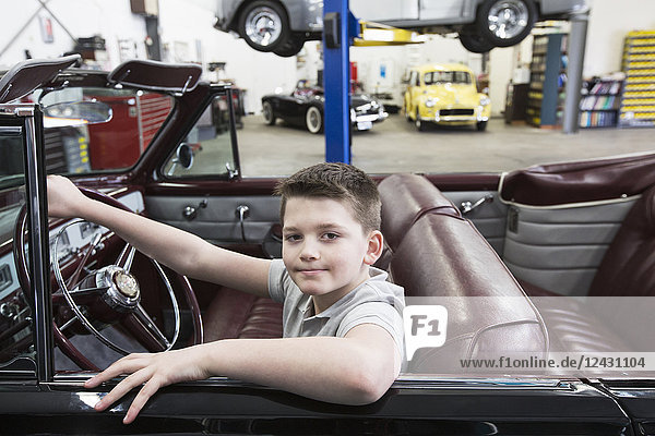 A young Caucasian boy at the wheel of an old convertible in his dad's classic car repair shop.