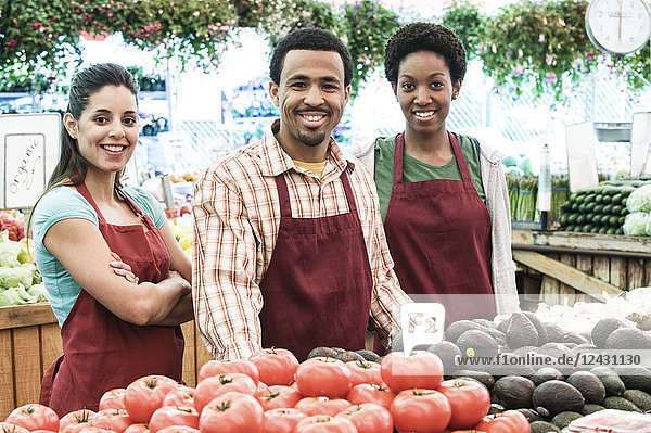 Mann und zwei Frauen in Schürzen stehen an einem Stand mit frischen Tomaten auf einem Obst- und Gemüsemarkt.