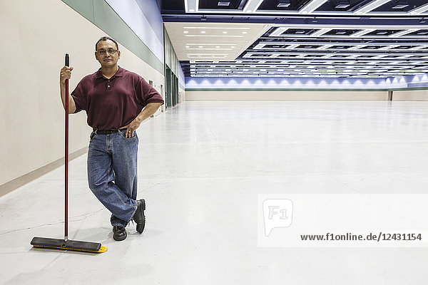 An Hispanic workman standing in a large interior space with a broom.