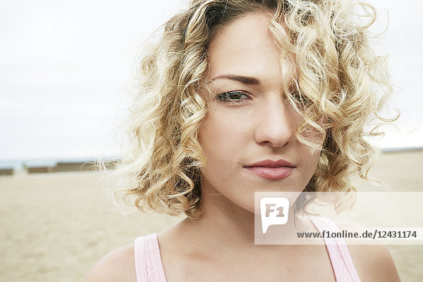 Porträt einer jungen Frau mit blonden Lockenhaaren  die am Sandstrand steht und in die Kamera schaut.
