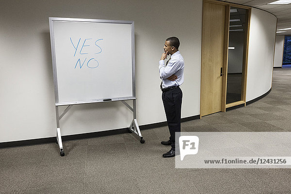 A black businessman thinking and standing in front of an office white board with the words Yes and No written on it.
