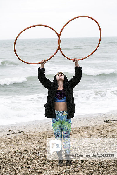 Junge Frau mit braunen Haaren und Dreadlocks  die an einem Sandstrand am Meer steht und zwei Hula-Hoop-Reifen balanciert.