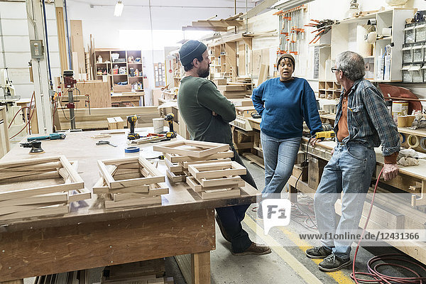A group of mixed race carpenters discussing a project at a work station in a large woodworking shop.