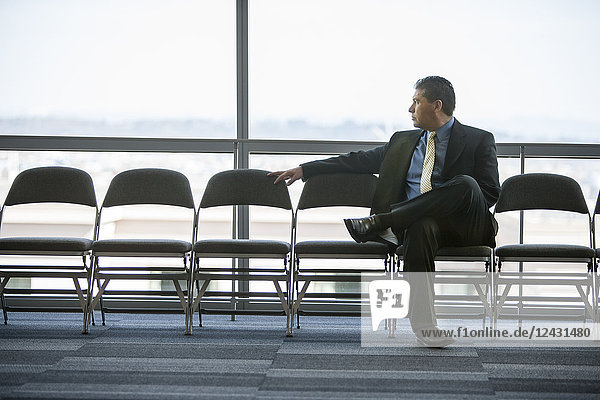 Hispanischer Geschäftsmann  der in einer Stuhlreihe vor einem Fenster in der Lobby eines Konferenzzentrums sitzt.