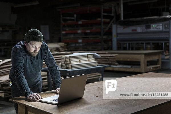 A Caucasian carpenter working on his lap top after hours in a large woodworking factory.