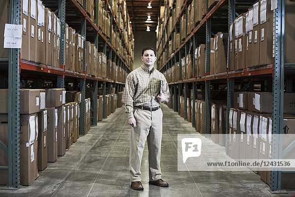 A Hispanic male warehouse worker standing next to racks of boxed products in a distribution warehouse.
