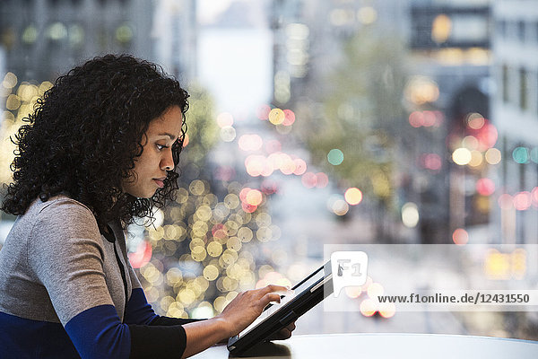 A view of a black businesswoman working on a notebook computer in front of an office window looking out on a city street scene just before dark.