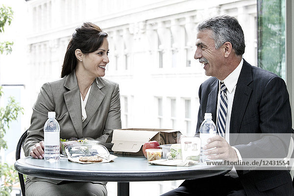 A Caucasian businesswoman and a middle eastern businessman having a business lunch in front of a convention centre lobby window.
