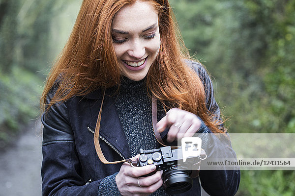 Lächelnde junge Frau mit langen roten Haaren  die einen Waldweg entlang läuft und mit einer Oldtimer-Kamera fotografiert.