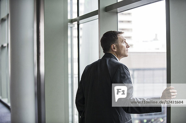 A Caucasian businessman standing next to and looking out of a large window in a convention centre.