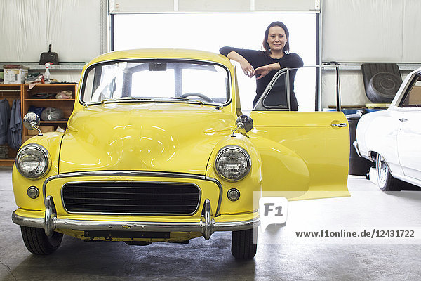 A Caucasian female stands next to her old car in a classic car repair shop.