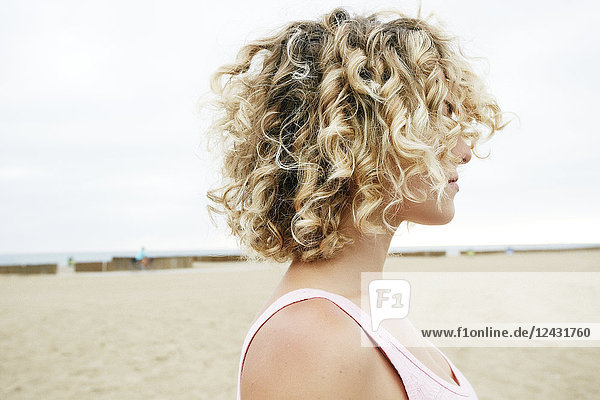Profilporträt einer jungen Frau mit blonden Lockenhaaren  die am Sandstrand steht.