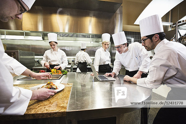 A crew of chef's working in a commercial kitchen  while several chefs wok on a laptop computer.