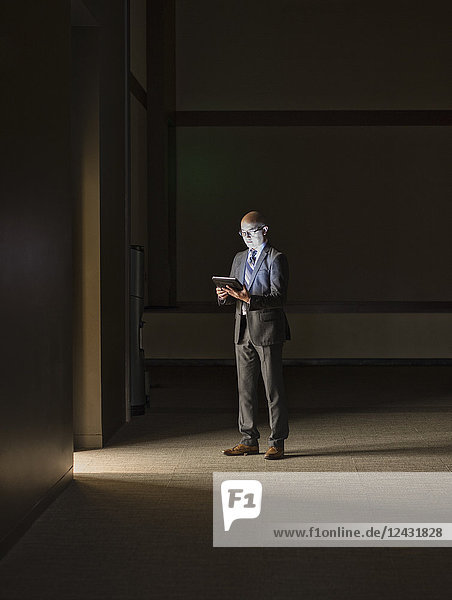 A mature Asian businessman standing in shadows in an office building  looking at a digital tablet.
