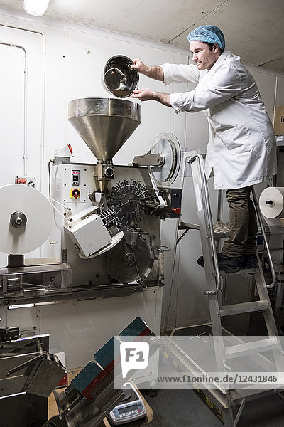 Man wearing white coat standing on a ladder  pouring tea leaves into large funnel of a machine.
