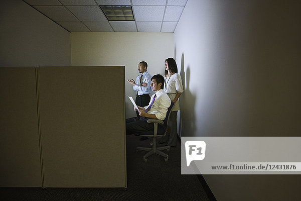 Eine gemischtrassige Gruppe von drei Geschäftsleuten  beleuchtet von einem Desktop-Computerbildschirm in einem kleinen Eckkabinenbüro.