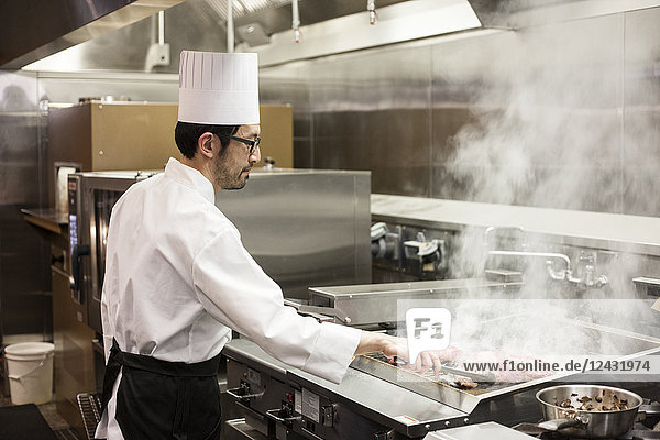 Ein asiatischer Küchenchef  der in einer Großküche mit frischem Fleisch auf dem Grill arbeitet