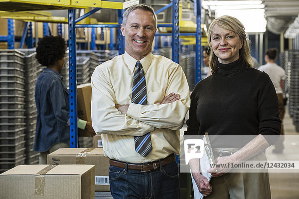 Portrait of a Caucasian male executive in a shirt and tie and a Caucasian female executive in sweater and slacks standing next to a motorized conveyor system in a large distribution warehouse.