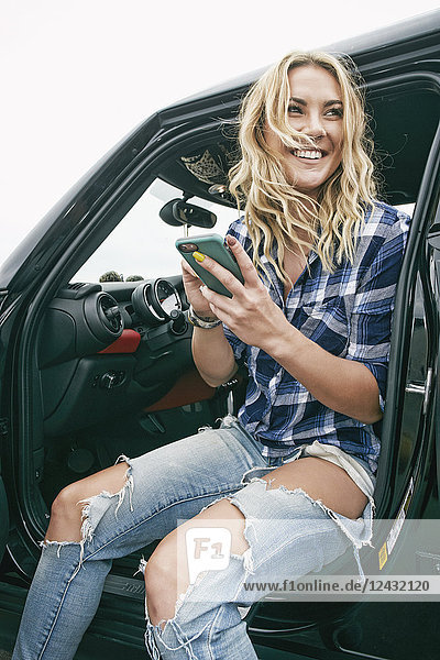Lächelnde Frau mit langen blonden Haaren  blau kariertem Hemd und zerrissenen Jeans  die im Auto sitzt und ihr Handy in der Hand hält.
