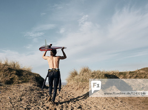 Mann  der mit einem auf dem Kopf balancierenden Surfbrett auf einem Pfad in den Sanddünen Richtung Meer läuft