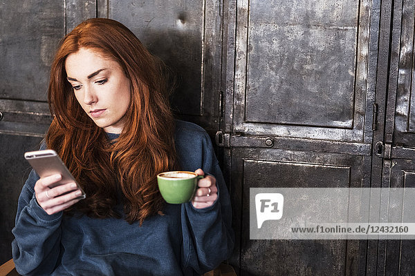 Junge Frau mit langen roten Haaren sitzt am Tisch  hält eine Tasse Kaffee in der Hand und überprüft ihr Mobiltelefon.