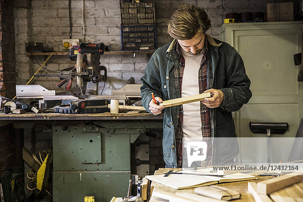 Mann arbeitet stehend an einer Werkbank in einer Holzbearbeitungswerkstatt und hält ein Stück Holz.