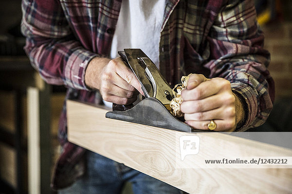 Nahaufnahme eines Mannes  der stehend in einer Holzwerkstatt arbeitet  mit einem Hobel auf einem Brett aus Holz.