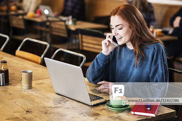 Smiling young woman with long red hair sitting at table  working on laptop computer  using mobile phone.