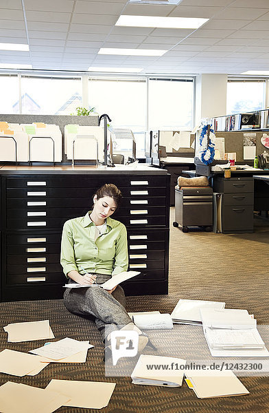 Eine junge kaukasische Geschäftsfrau sitzt auf dem Boden ihres Büros und geht Papierkram durch.