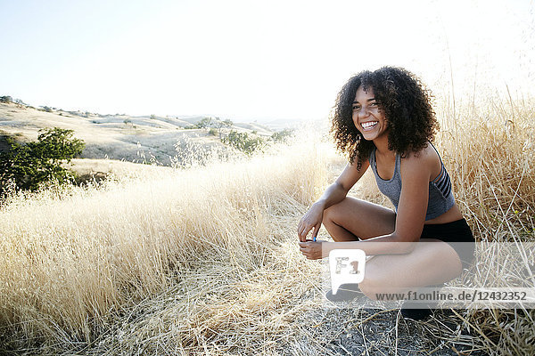 Lächelnde junge Frau mit lockigem braunen Haar im Stadtpark  kniend auf einem Feldweg.