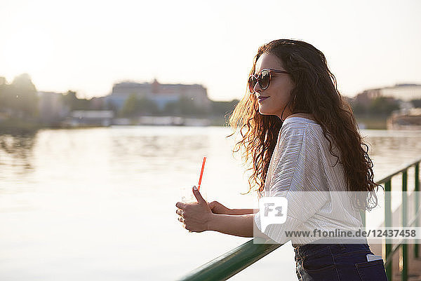 Smiling young woman with a takeaway drink enjoying the sunset at the riverside