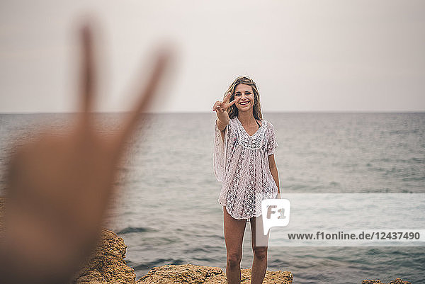Portrait of happy young woman standing on a rock at the sea making peace sign