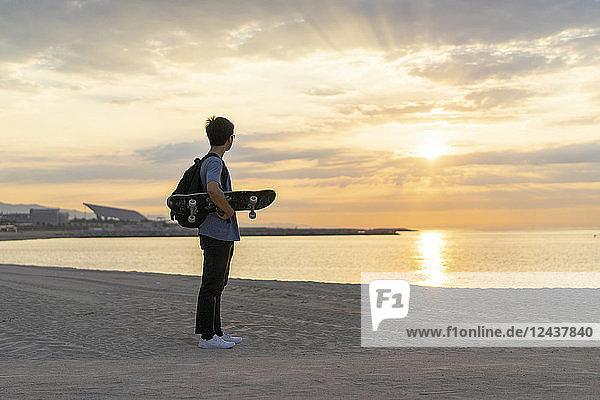 Young Chinese man with skateboard standing at the beach at sunrise