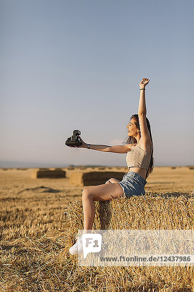 Young woman sitting on straw bale taking selfie with instant camera