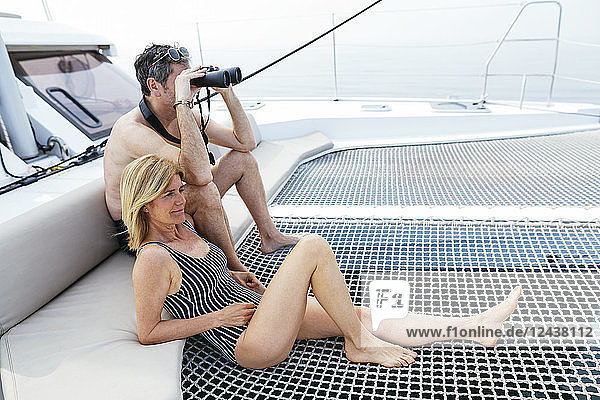 Mature couple sitting on catamaran trampoline  using binoculars