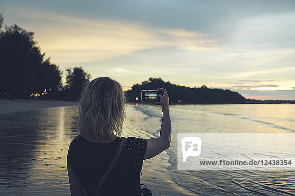 Thailand  Khao Lak  back view of woman taking photo with cell phone on the beach at sunset