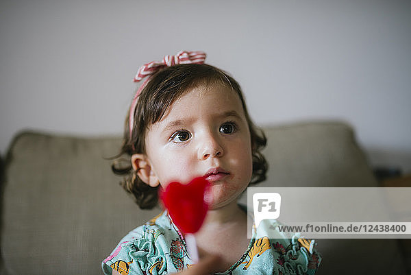 Cute baby girl eating a heart shaped lollipop at home