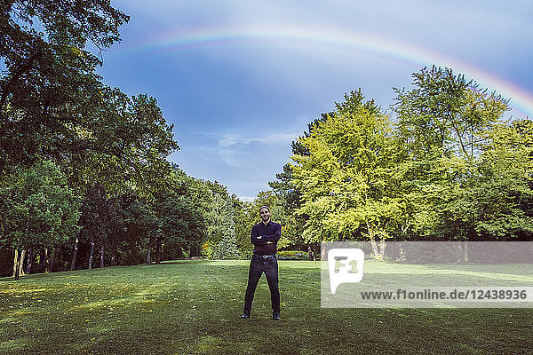 Young businessman standing in park under a rainbow  with arms crossed