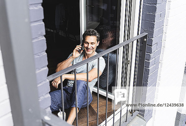 Smiling man in pyjama at home on cell phone at balcony door