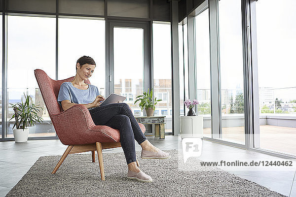 Woman sitting in armchair at home using tablet