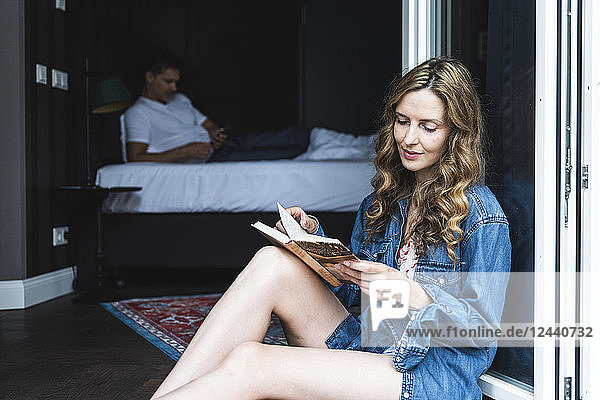 Woman reading book sitting at French window with man in background